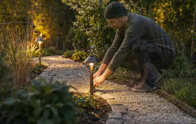 Person richtet eine niedrige warm leuchtende Gartenlampe am Kiesweg aus; Thema Gartenhaus aus Kunststoff, Schwerpunkt Nachhaltigkeit; Umsetzung.