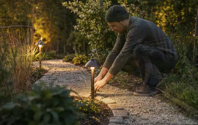 Person richtet eine niedrige warm leuchtende Gartenlampe am Kiesweg aus; Thema Gartenhaus aus Kunststoff, Schwerpunkt Pflege; Ausgangslage.