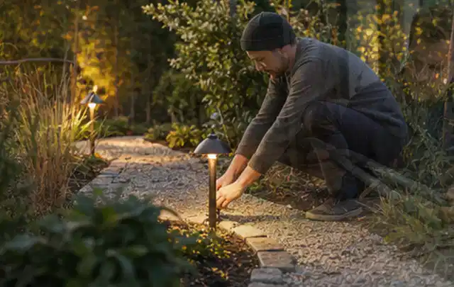 Person richtet eine niedrige warm leuchtende Gartenlampe am Kiesweg aus; Thema Gartenhaus aus Kunststoff, Schwerpunkt Planung; Ausgangslage.