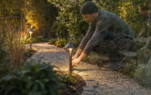 Person richtet eine niedrige warm leuchtende Gartenlampe am Kiesweg aus; Thema Gartenhaus aus Kunststoff, Schwerpunkt Standort; Ausgangslage.