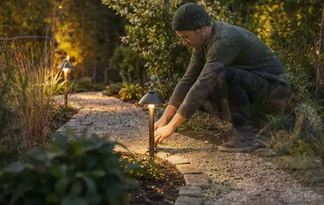 Person richtet eine niedrige warm leuchtende Gartenlampe am Kiesweg aus; Thema Gartenhaus aus Metall, Schwerpunkt Baurecht; Praxisalltag.