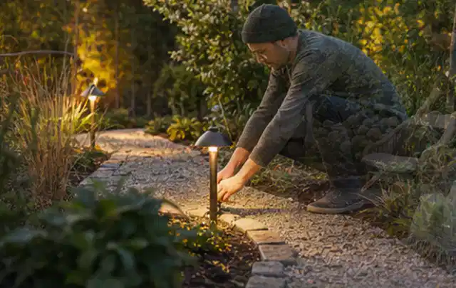 Person richtet eine niedrige warm leuchtende Gartenlampe am Kiesweg aus; Thema Gartenhaus aus Metall, Schwerpunkt Fundament; Umsetzung.