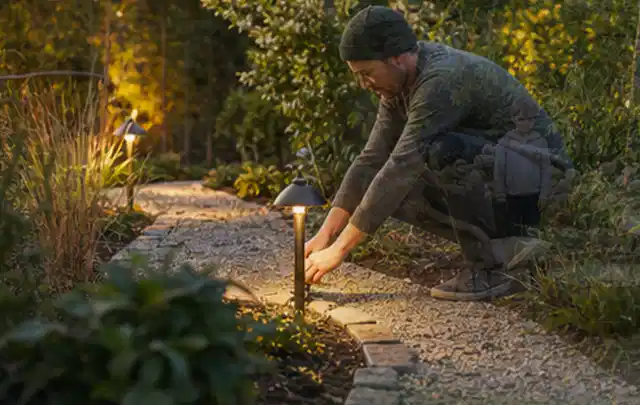 Person richtet eine niedrige warm leuchtende Gartenlampe am Kiesweg aus; Thema Gartenhaus aus Metall, Schwerpunkt Gestaltung; Umsetzung.