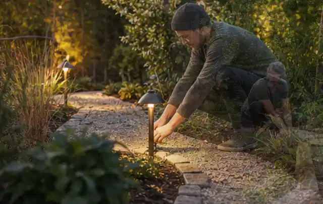Person richtet eine niedrige warm leuchtende Gartenlampe am Kiesweg aus; Thema Gartenhaus aus Metall, Schwerpunkt Kosten; Praxisalltag.