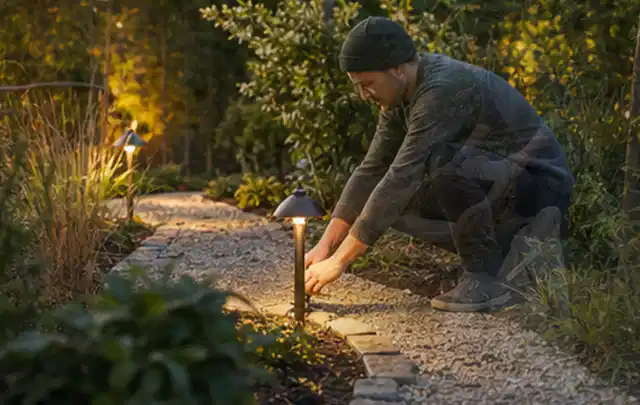 Person richtet eine niedrige warm leuchtende Gartenlampe am Kiesweg aus; Thema Gartenhaus aus Metall, Schwerpunkt Nachhaltigkeit; Kontrolle.