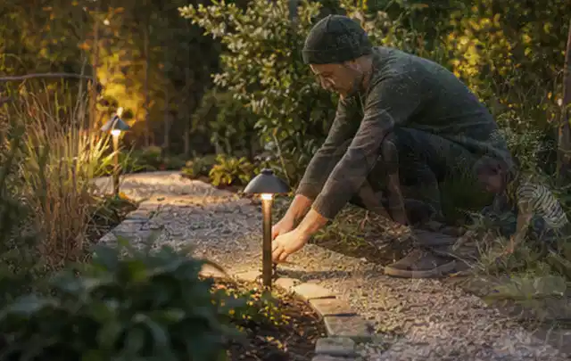 Person richtet eine niedrige warm leuchtende Gartenlampe am Kiesweg aus; Thema Gartenhaus aus Metall, Schwerpunkt Winterschutz; Umsetzung.