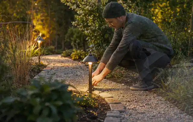 Person richtet eine niedrige warm leuchtende Gartenlampe am Kiesweg aus; Thema Gartenhaus Dach, Schwerpunkt Aufbau; Kontrolle.
