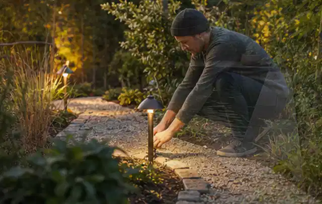 Person richtet eine niedrige warm leuchtende Gartenlampe am Kiesweg aus; Thema Gartenhaus Dach, Schwerpunkt Dimensionierung; Kontrolle.