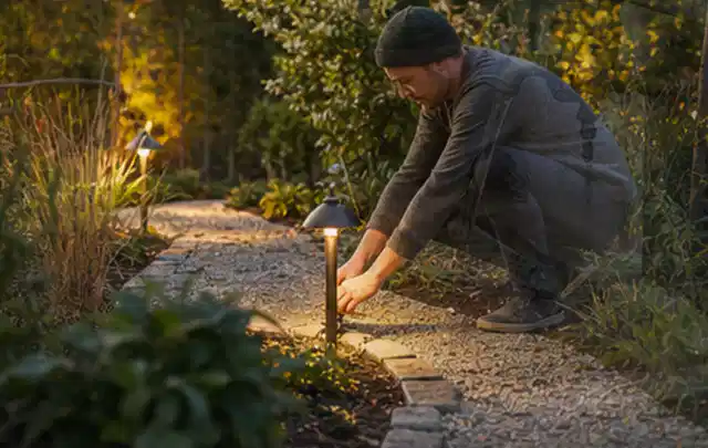 Person richtet eine niedrige warm leuchtende Gartenlampe am Kiesweg aus; Thema Gartenhaus Dach, Schwerpunkt DIY; Umsetzung.