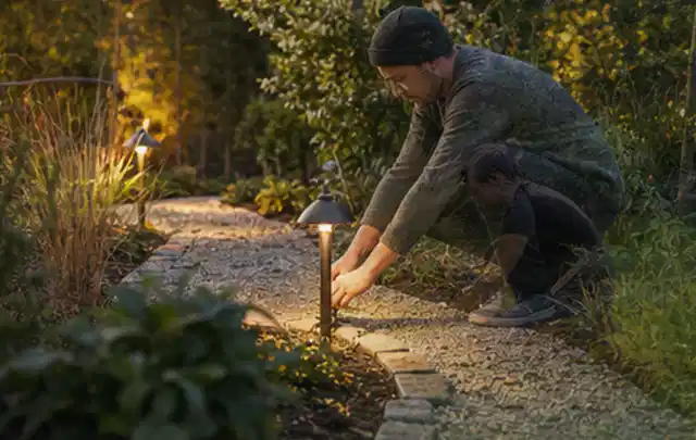 Person richtet eine niedrige warm leuchtende Gartenlampe am Kiesweg aus; Thema Gartenhaus Dach, Schwerpunkt Kosten; Ausgangslage.