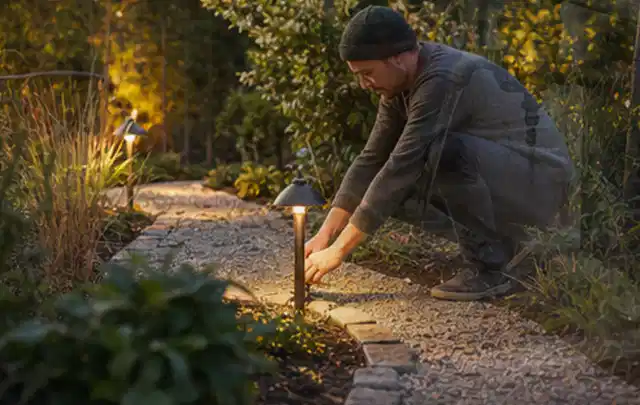 Person richtet eine niedrige warm leuchtende Gartenlampe am Kiesweg aus; Thema Gartenhaus Dach, Schwerpunkt Materialwahl; Kontrolle.