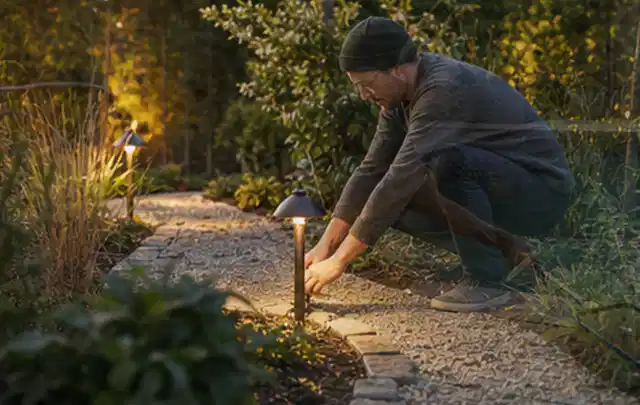 Person richtet eine niedrige warm leuchtende Gartenlampe am Kiesweg aus; Thema Gartenhaus Fenster, Schwerpunkt Dimensionierung; Umsetzung.
