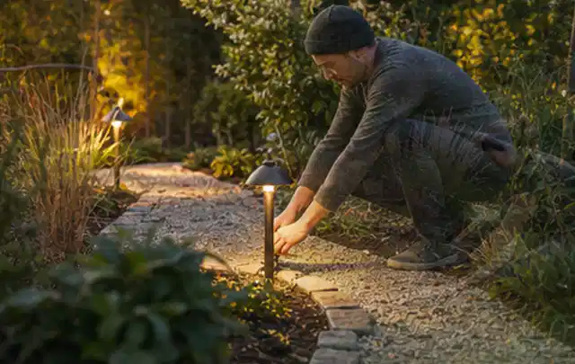 Person richtet eine niedrige warm leuchtende Gartenlampe am Kiesweg aus; Thema Gartenhaus Fenster, Schwerpunkt Fehlerhilfe; Ergebnis.