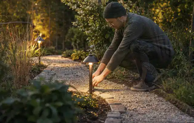 Person richtet eine niedrige warm leuchtende Gartenlampe am Kiesweg aus; Thema Gartenhaus Fenster, Schwerpunkt Pflege; Ausgangslage.