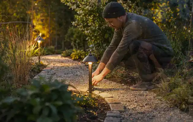 Person richtet eine niedrige warm leuchtende Gartenlampe am Kiesweg aus; Thema Gartenhaus Fundament, Schwerpunkt DIY; Umsetzung.