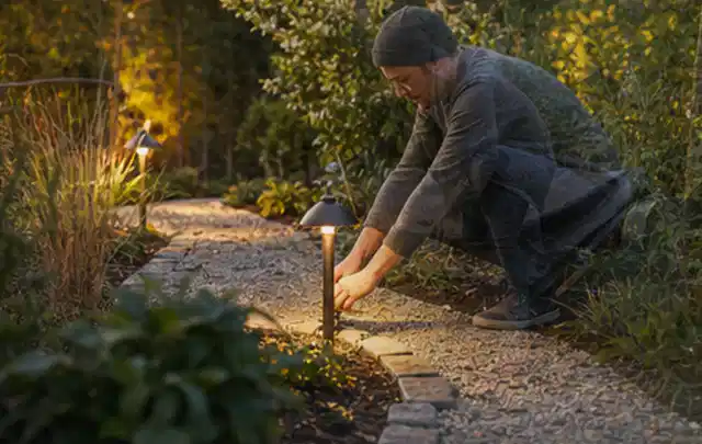 Person richtet eine niedrige warm leuchtende Gartenlampe am Kiesweg aus; Thema Gartenhaus Fundament, Schwerpunkt Fehlerhilfe; Ergebnis.