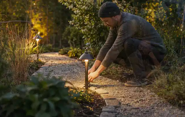 Person richtet eine niedrige warm leuchtende Gartenlampe am Kiesweg aus; Thema Gartenhaus Fundament, Schwerpunkt Gärten mit kleiner Fläche; Umsetzung.