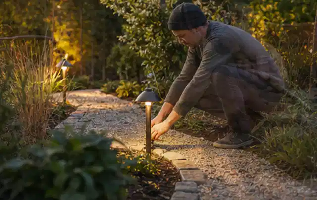 Person richtet eine niedrige warm leuchtende Gartenlampe am Kiesweg aus; Thema Gartenhaus Fundament, Schwerpunkt Gestaltung; Praxisalltag.