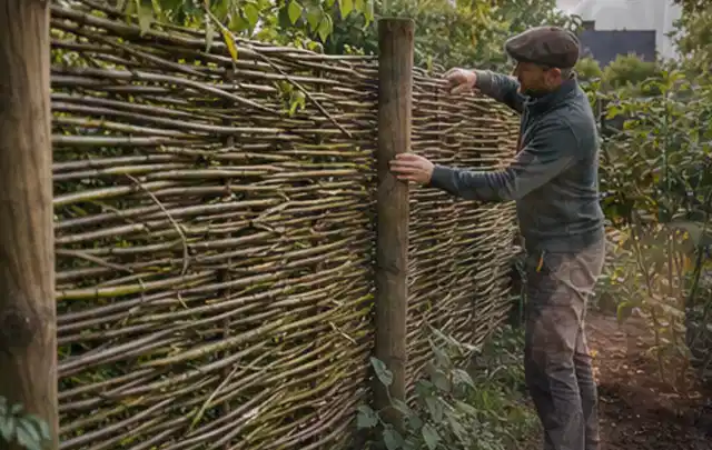 Person befestigt geflochtene Weidenruten an stabilen Holzpfosten im Garten; Thema Gartenhaus Fundament, Schwerpunkt Materialwahl; Umsetzung.
