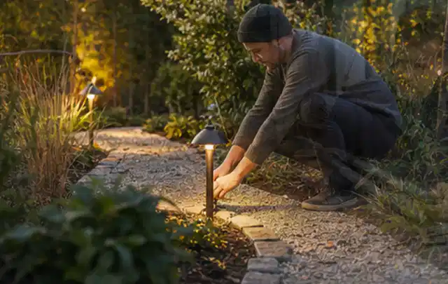Person richtet eine niedrige warm leuchtende Gartenlampe am Kiesweg aus; Thema Gartenhaus Fundament, Schwerpunkt Materialwahl; Praxisalltag.