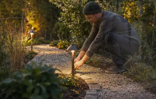 Person richtet eine niedrige warm leuchtende Gartenlampe am Kiesweg aus; Thema Gartenhaus Fundament, Schwerpunkt Planung; Kontrolle.