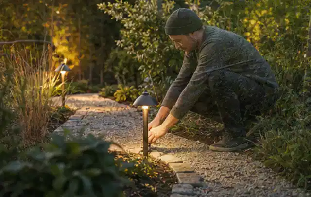 Person richtet eine niedrige warm leuchtende Gartenlampe am Kiesweg aus; Thema Gartenhaus Genehmigung, Schwerpunkt Gartenhäuser; Ergebnis.