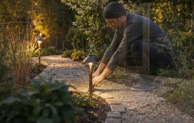 Person richtet eine niedrige warm leuchtende Gartenlampe am Kiesweg aus; Thema Gartenlounges, Schwerpunkt Vergleich; Ergebnis.