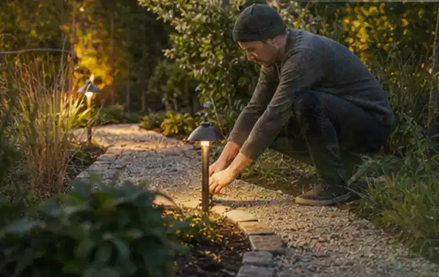Person richtet eine niedrige warm leuchtende Gartenlampe am Kiesweg aus; Thema Gartenlounges, Schwerpunkt Wartung; Ergebnis.