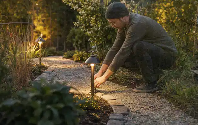 Person richtet eine niedrige warm leuchtende Gartenlampe am Kiesweg aus; Thema Gartenlounges, Schwerpunkt Winterlager; Kontrolle.