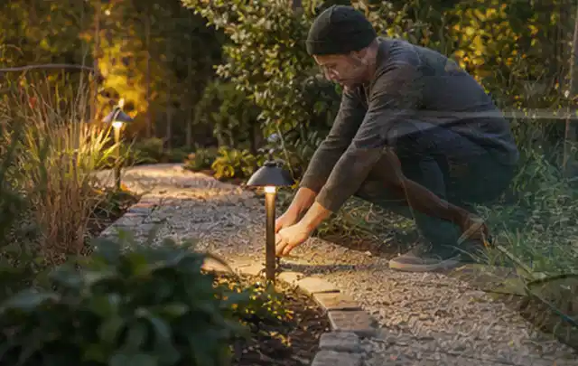 Person richtet eine niedrige warm leuchtende Gartenlampe am Kiesweg aus; Thema Gartenneuanlage, Schwerpunkt Fehlerhilfe; Praxisalltag.