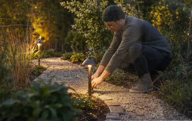 Person richtet eine niedrige warm leuchtende Gartenlampe am Kiesweg aus; Thema Gartenneuanlage, Schwerpunkt Materialwahl; Praxisalltag.