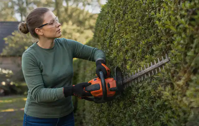 Person schneidet eine dichte Gartenhecke mit Schutzbrille und sicherem Stand; Thema Gartenpumpen, Schwerpunkt Sicherheit; Kontrolle.