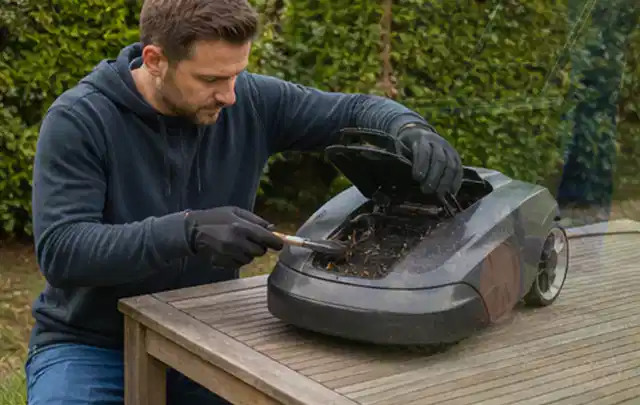 Person wartet einen geöffneten Mähroboter auf einem stabilen Gartentisch; Thema Gartenscheren, Schwerpunkt Reinigung; Kontrolle.