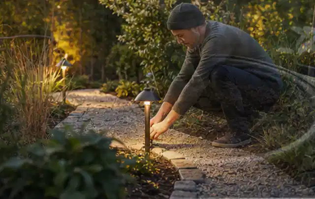 Person richtet eine niedrige warm leuchtende Gartenlampe am Kiesweg aus; Thema Gartenstühle, Schwerpunkt Anwendung; Kontrolle.