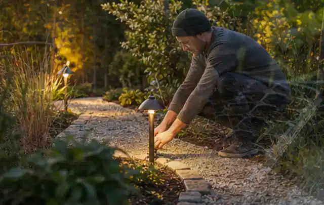 Person richtet eine niedrige warm leuchtende Gartenlampe am Kiesweg aus; Thema Gartenstühle, Schwerpunkt Checkliste; Kontrolle.