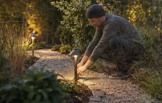 Person richtet eine niedrige warm leuchtende Gartenlampe am Kiesweg aus; Thema Gartenstühle, Schwerpunkt Ersatzteile; Ausgangslage.