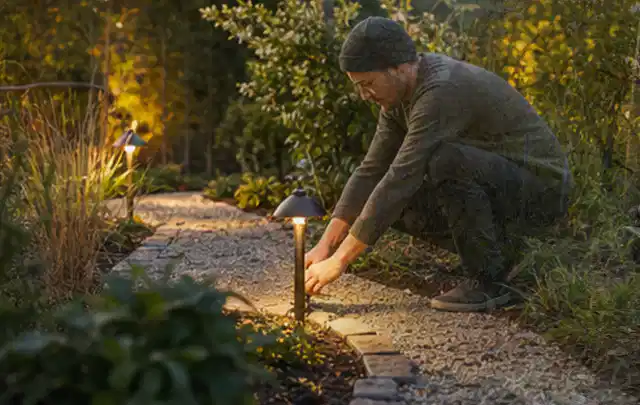 Person richtet eine niedrige warm leuchtende Gartenlampe am Kiesweg aus; Thema Gartenstühle, Schwerpunkt Gärten mit großer Fläche; Umsetzung.