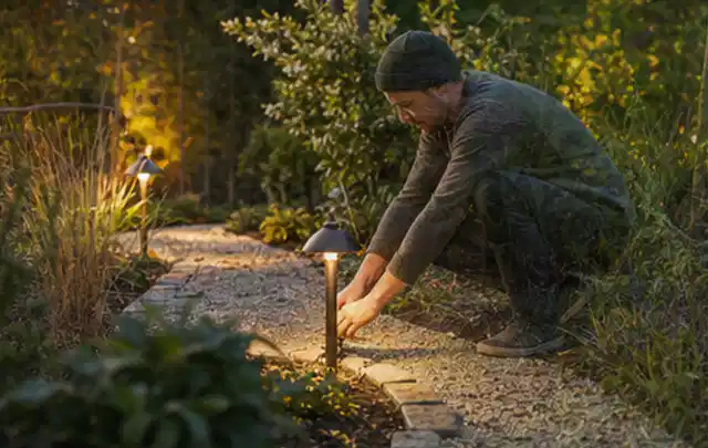 Person richtet eine niedrige warm leuchtende Gartenlampe am Kiesweg aus; Thema Gartenstühle, Schwerpunkt Vergleich; Ausgangslage.