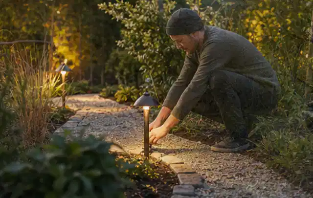 Person richtet eine niedrige warm leuchtende Gartenlampe am Kiesweg aus; Thema Gartentische, Schwerpunkt Checkliste; Ausgangslage.