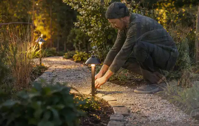 Person richtet eine niedrige warm leuchtende Gartenlampe am Kiesweg aus; Thema Gartentische, Schwerpunkt Dimensionierung; Umsetzung.