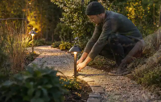 Person richtet eine niedrige warm leuchtende Gartenlampe am Kiesweg aus; Thema Gartentische, Schwerpunkt Gärten mit großer Fläche; Kontrolle.