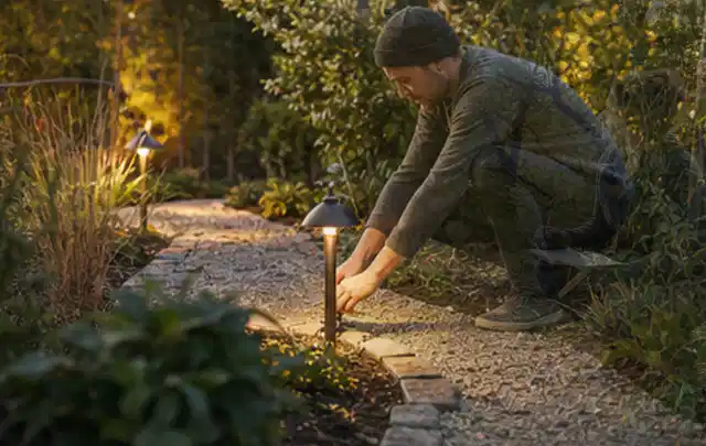 Person richtet eine niedrige warm leuchtende Gartenlampe am Kiesweg aus; Thema Gartentische, Schwerpunkt Gärten mit kleiner Fläche; Praxisalltag.