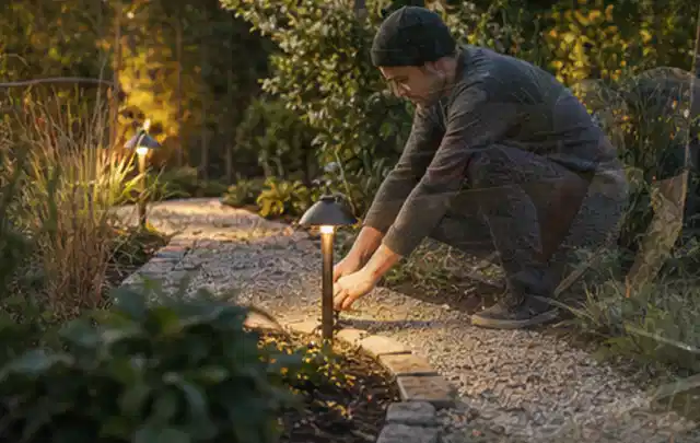 Person richtet eine niedrige warm leuchtende Gartenlampe am Kiesweg aus; Thema Gartentische, Schwerpunkt Nachhaltigkeit; Kontrolle.
