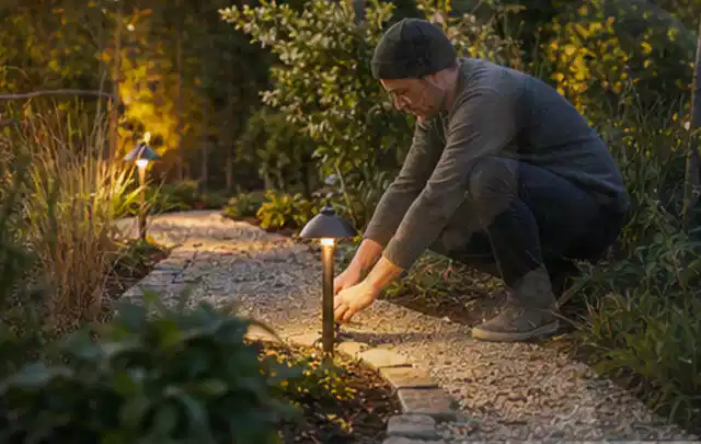 Person richtet eine niedrige warm leuchtende Gartenlampe am Kiesweg aus; Thema Gartentische, Schwerpunkt Reinigung; Ergebnis.