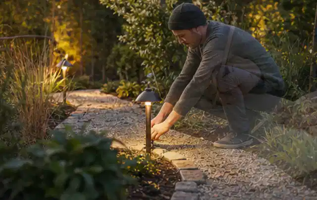 Person richtet eine niedrige warm leuchtende Gartenlampe am Kiesweg aus; Thema Gartentische, Schwerpunkt Reparatur; Ausgangslage.