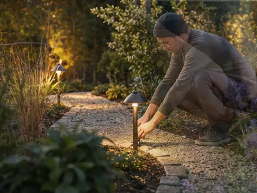Person richtet eine niedrige warm leuchtende Gartenlampe am Kiesweg aus; Thema Gartentische, Schwerpunkt Sicherheit; Titelbild.