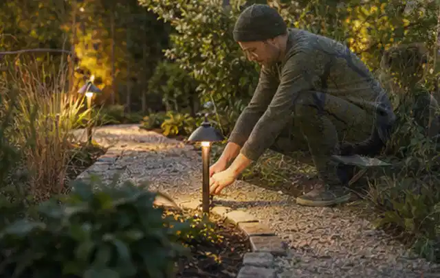 Person richtet eine niedrige warm leuchtende Gartenlampe am Kiesweg aus; Thema Gartentische, Schwerpunkt Vergleich; Umsetzung.