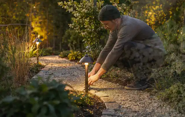 Person richtet eine niedrige warm leuchtende Gartenlampe am Kiesweg aus; Thema Gartentische, Schwerpunkt Wartung; Praxisalltag.