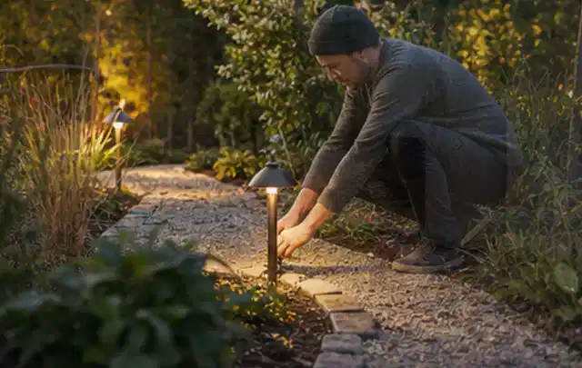Person richtet eine niedrige warm leuchtende Gartenlampe am Kiesweg aus; Thema Gartenwerkstatt, Schwerpunkt Fehlerhilfe; Umsetzung.