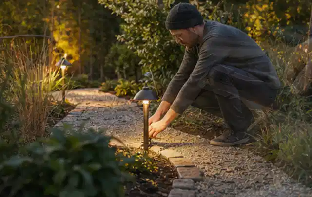 Person richtet eine niedrige warm leuchtende Gartenlampe am Kiesweg aus; Thema Gartenwerkstatt, Schwerpunkt Gärten mit kleiner Fläche; Ergebnis.